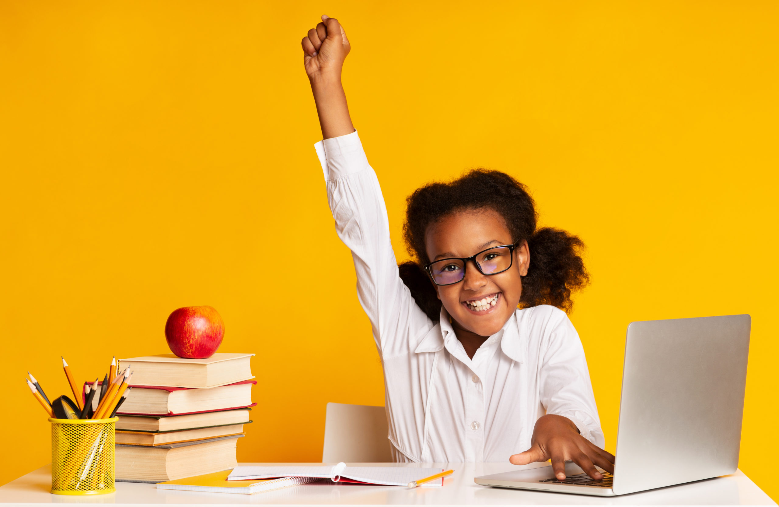 A girl child at a desk with books, an apple, and a laptop, raising one hand